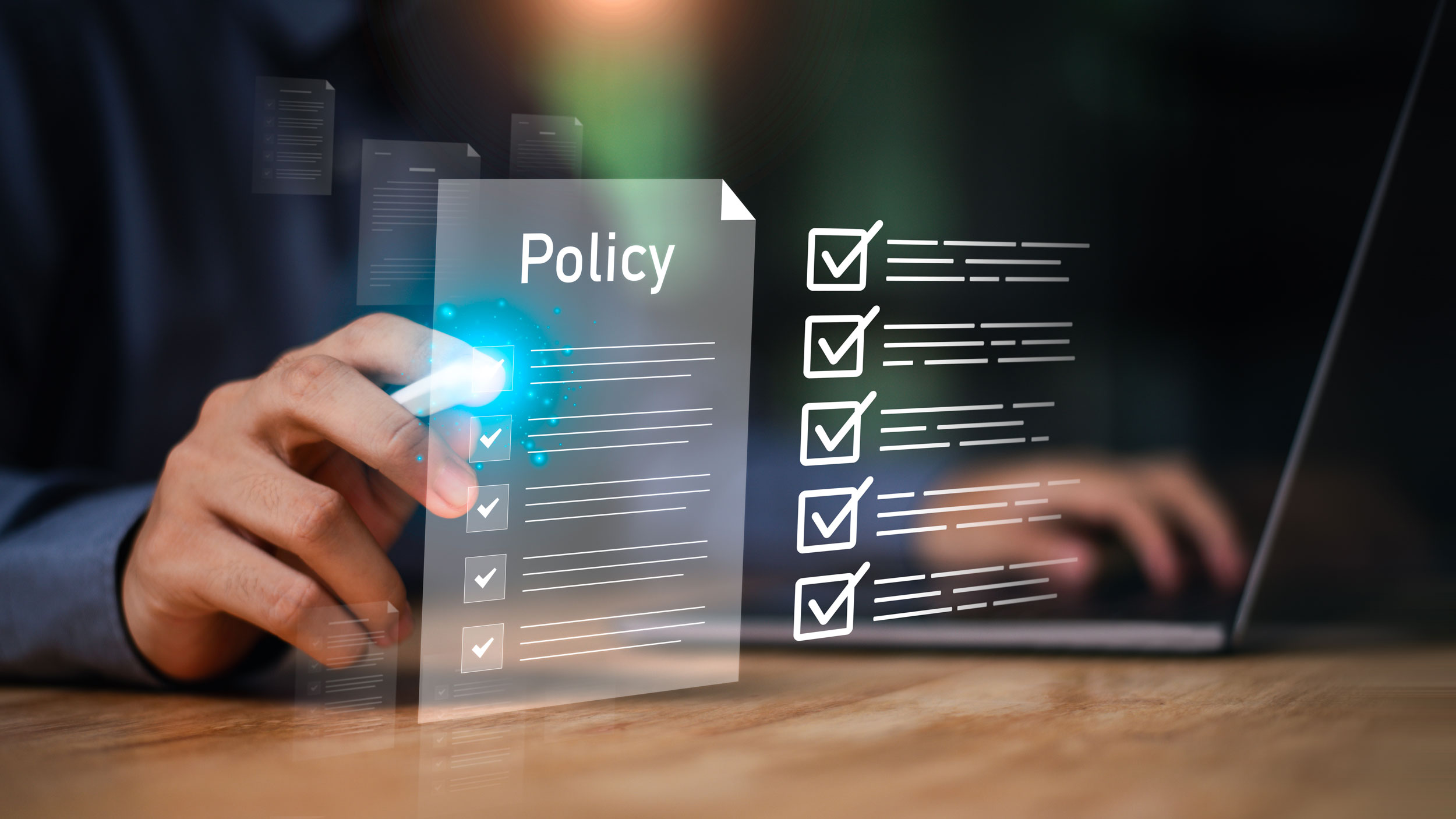 Ein Mann zeigt mit einem Stift auf ein Hologramm mit der Aufschrift "Policy" und einer abgehakten Checkliste. Im Hintergrund sieht man einen Laptop.