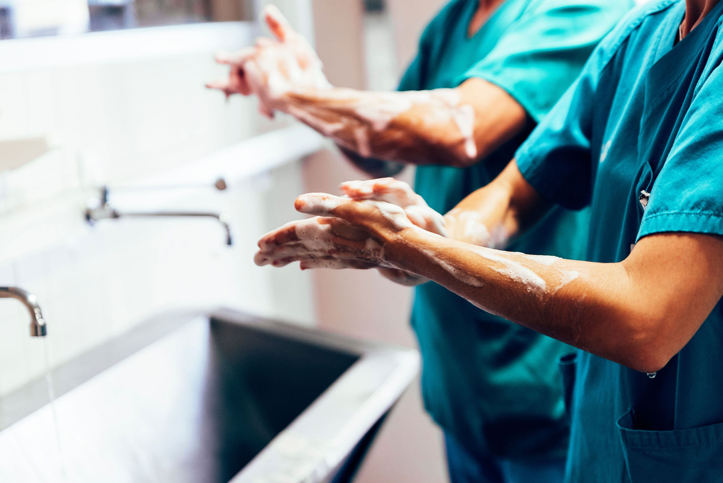 Zwei Personen in blauer Krankenhauskleidung waschen sich mit Seife die Hände und Unterarme. Sie stehen vor einem langen, tiefen Waschbecken.