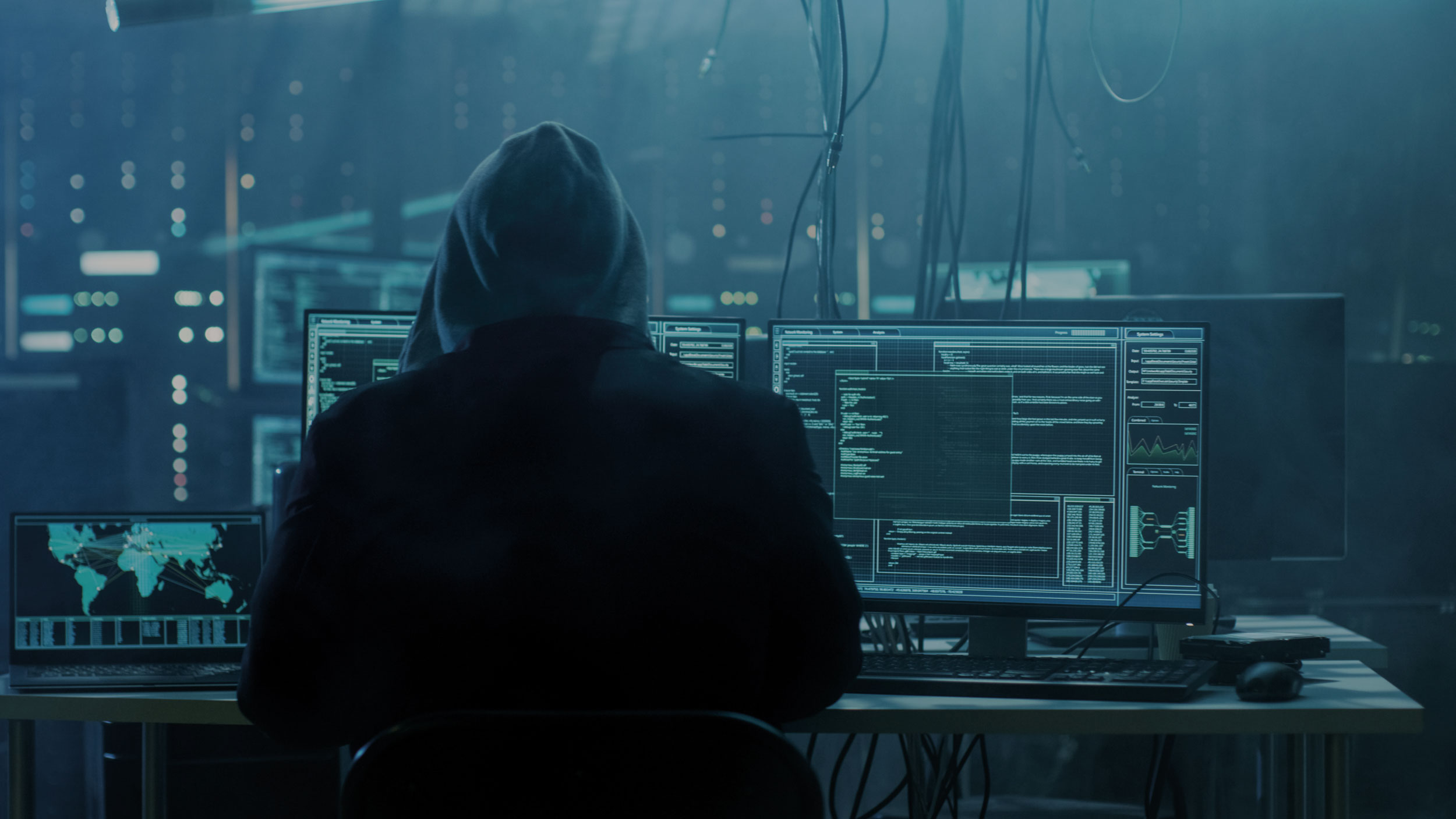Ein Hacker sitzt in einem düsteren Raum am PC. Im Hintergrund sieht man Server, Kabel und Bildschirme.
