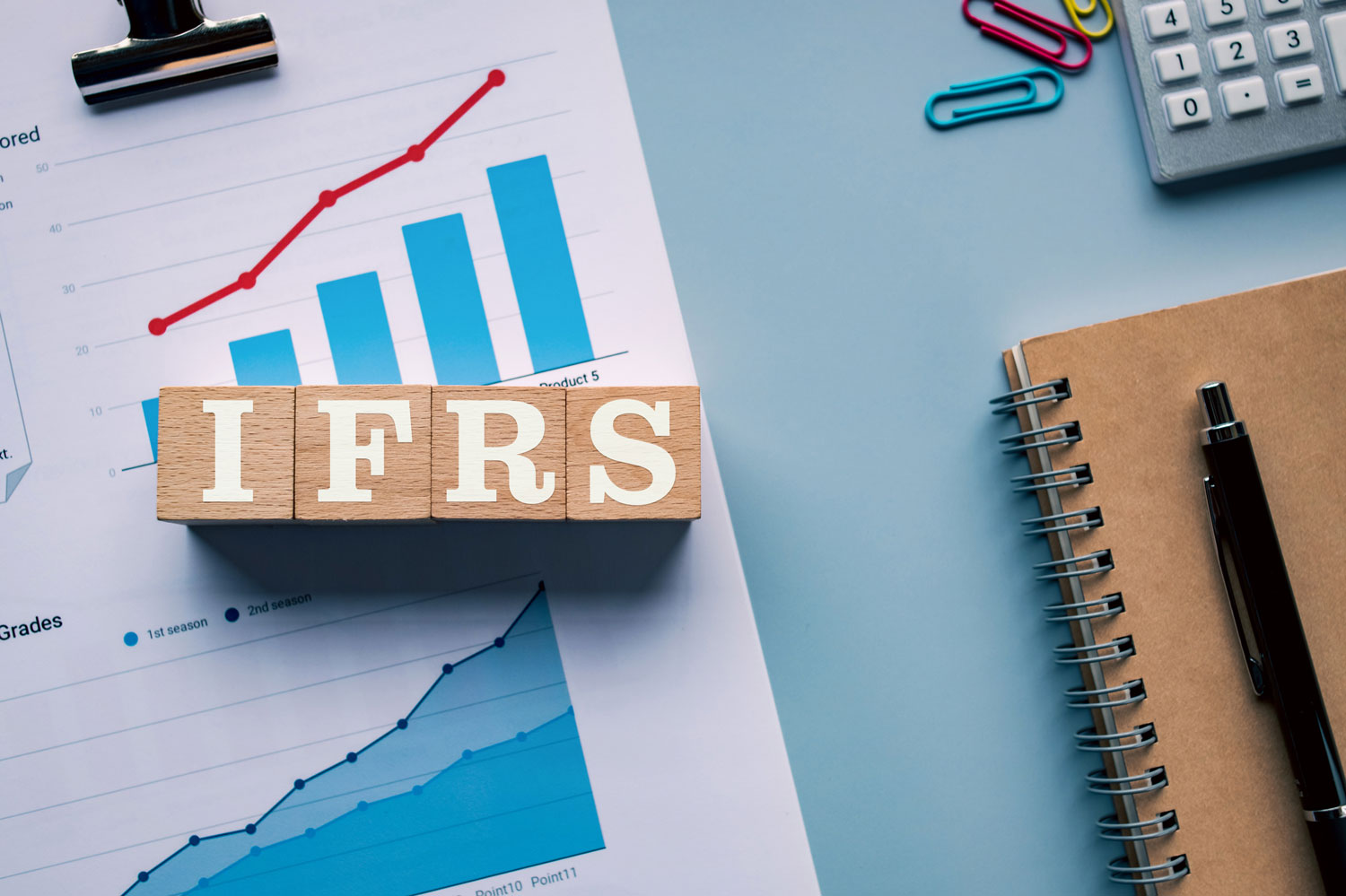Schreibtisch mit Taschenrechner, Büroklammern, Notizbuch & Kugelschreiber, Analysen sowie Würfel mit IFRS Schriftzug