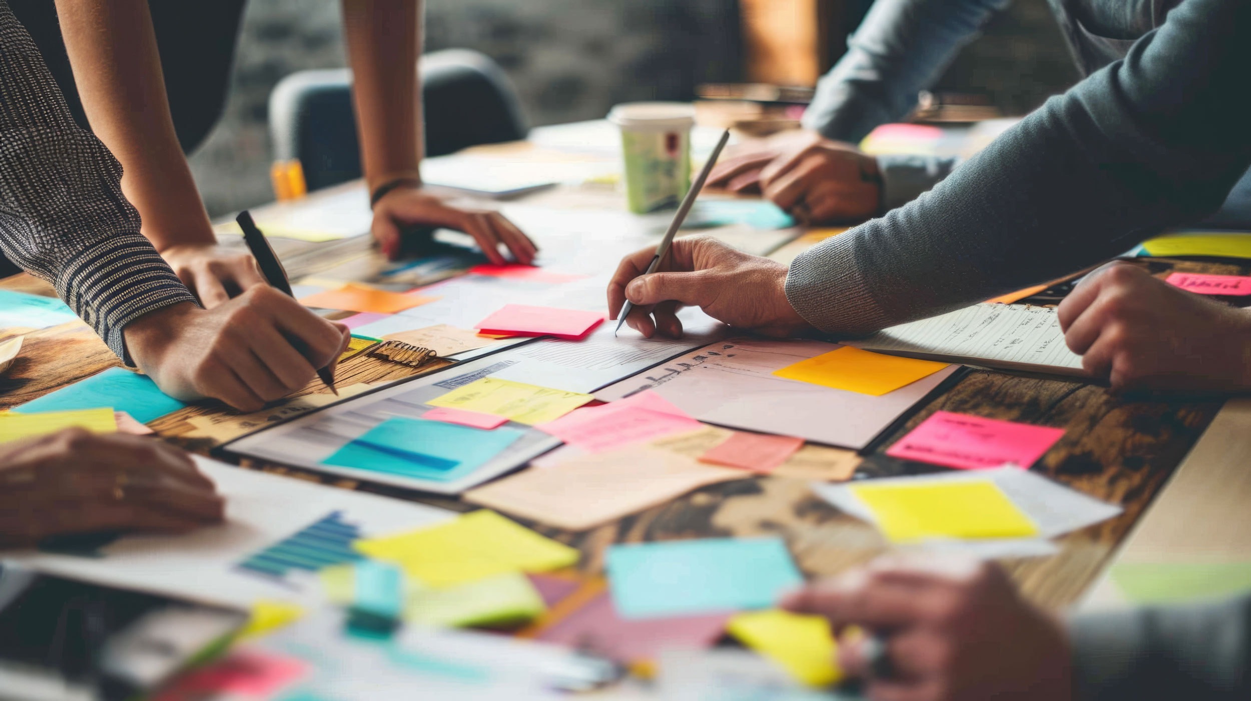 Menschen arbeiten an einem Tisch gemeinsam. Auf dem Tisch liegen Dokumente, Diagramm und bunte Post-its.