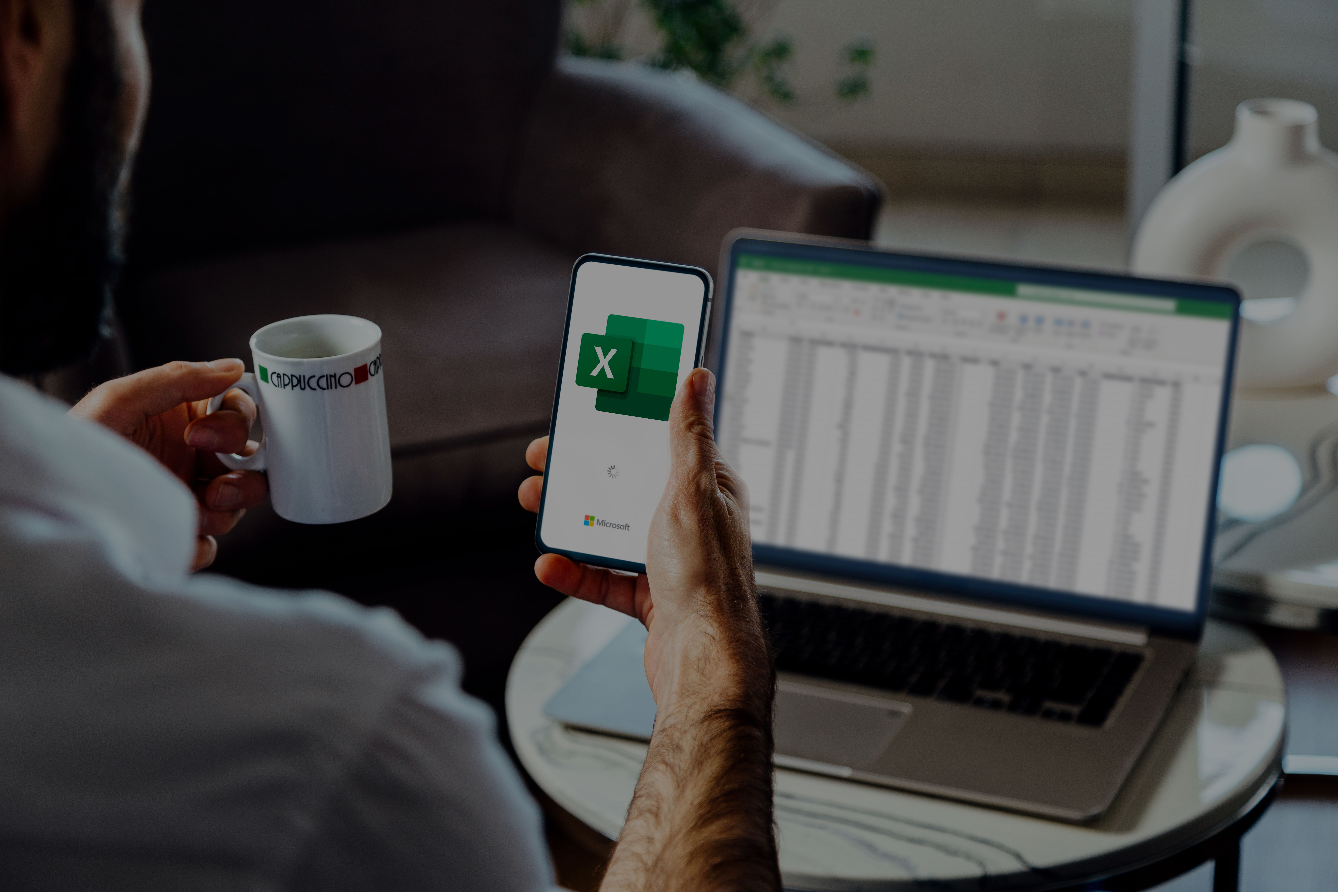 Ein Mann sitzt vor seinem Laptop und hält sein Handy in der Hand. Auf beiden Geräten verwendet er Microsoft Excel. In der anderen Hand hält er einen genüsslichen Kaffee.  