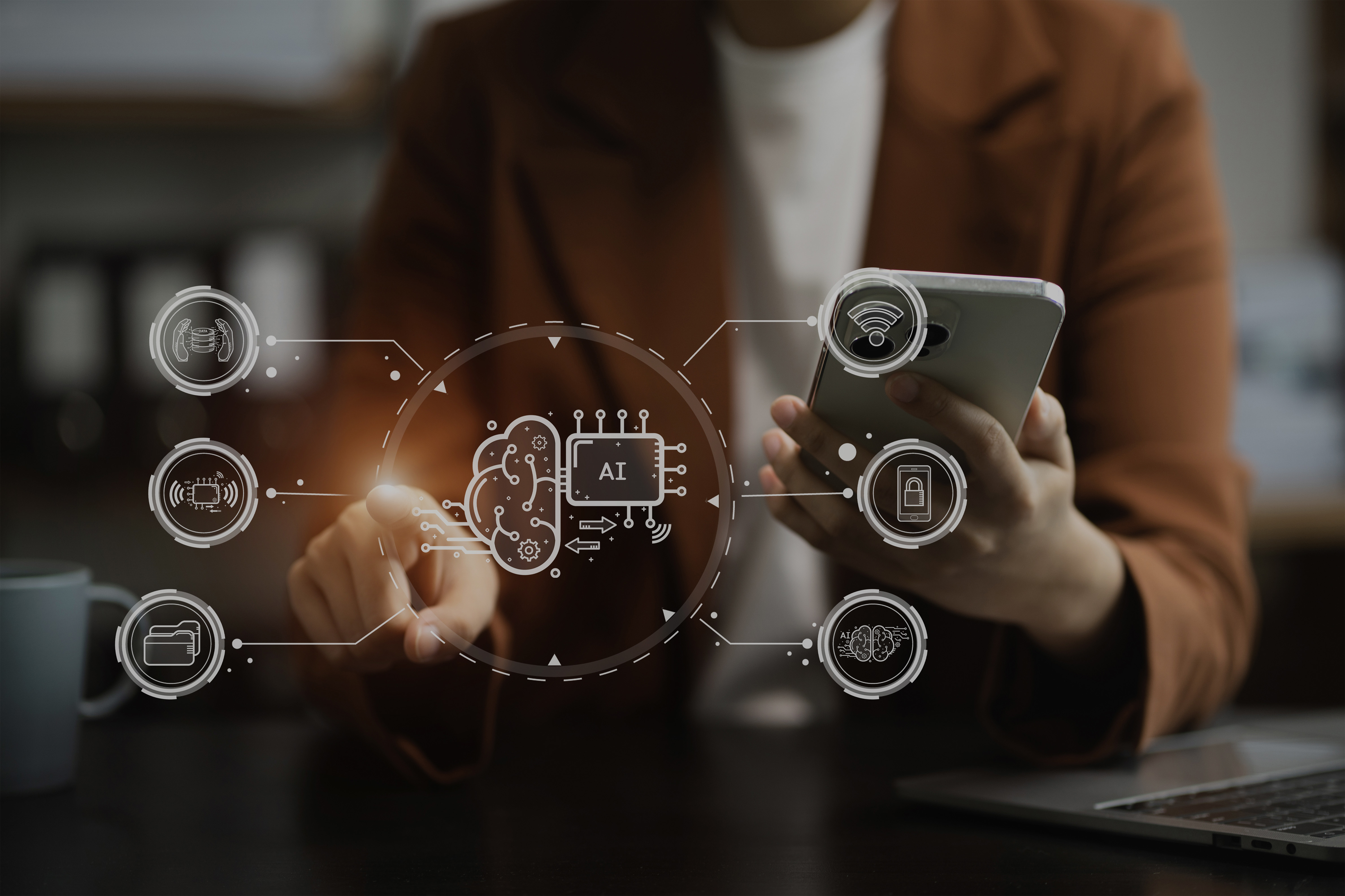 Im Vordergrund ist ein Hologramm mit verschiedenen Computer und KI Icons, im Hintergrund sitzt ein Mensch mit Handy in der der Hand uns zeigt mit der anderen Hand auf die Icons. Am Tisch im Vordergrund steht ein Laptop.