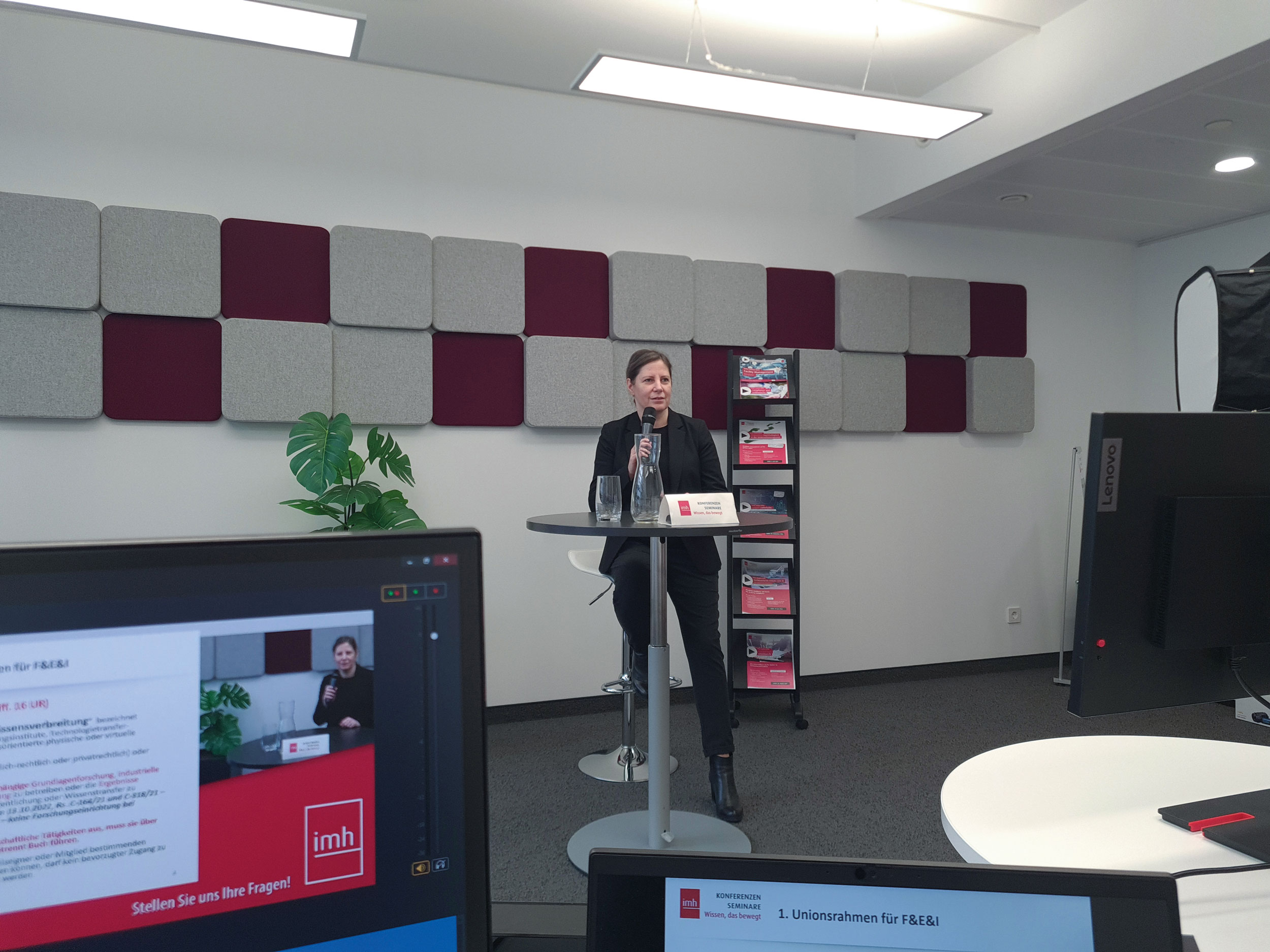 Im Vordergrund sieht man 2 Computerbildschirme. Hinterbei steht eine Frau bei einem Stehtisch und trägt für ein Webinar vor. 