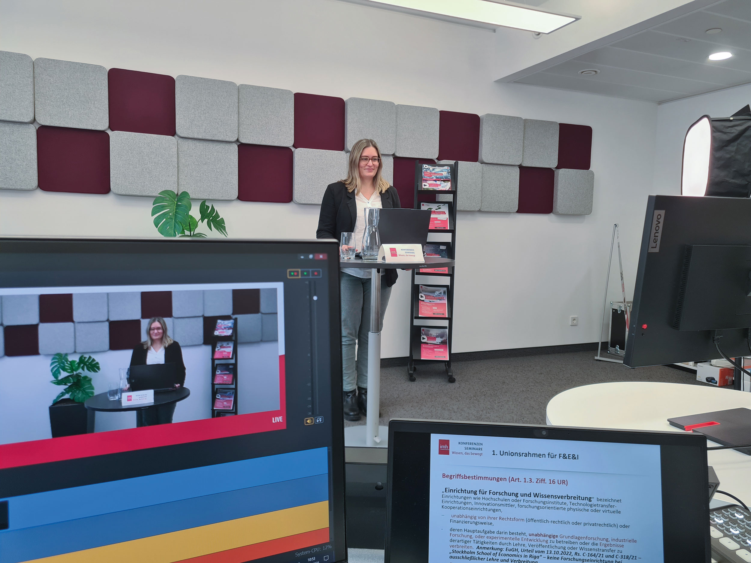 Im Vordergrund sieht man 2 Computerbildschirme. Hinterbei steht eine Frau bei einem Stehtisch und trägt für ein Webinar vor. 