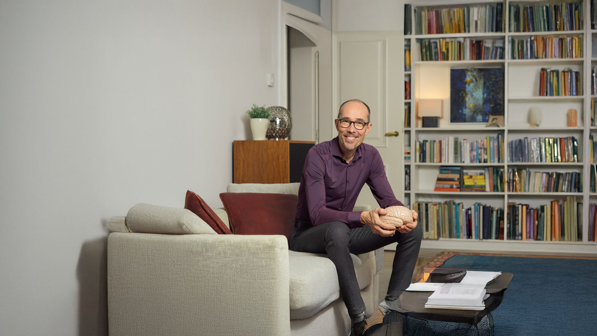 Bernd Hufnagel sitzt lächelnd auf der Couch und hält ein Modell von einem Gehirn.