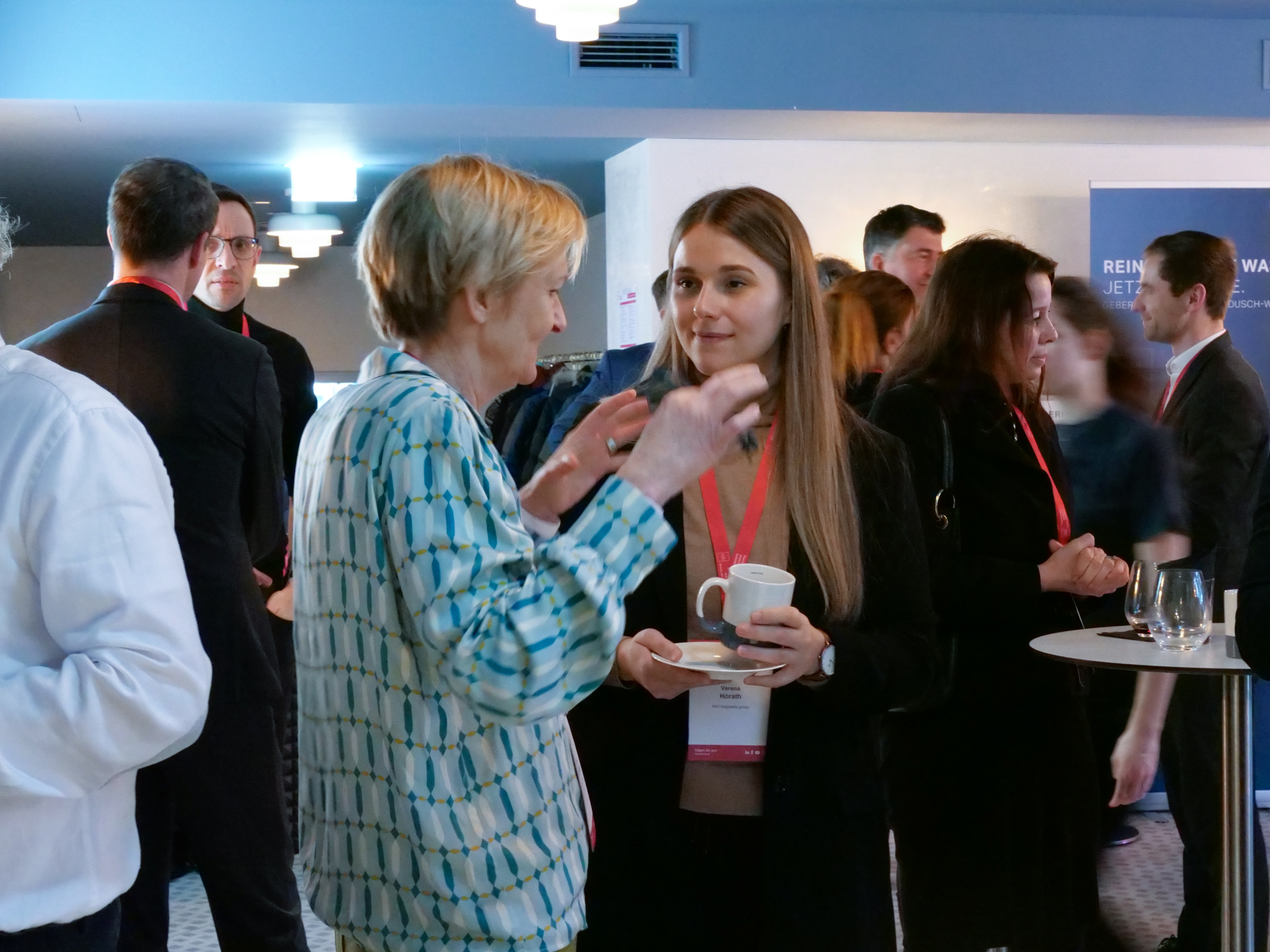 Zwei Frauen unterhalten sich. Eine hat eine Tasse in der Hand, im Hintergrund mehrere Menschen im Gespräch.