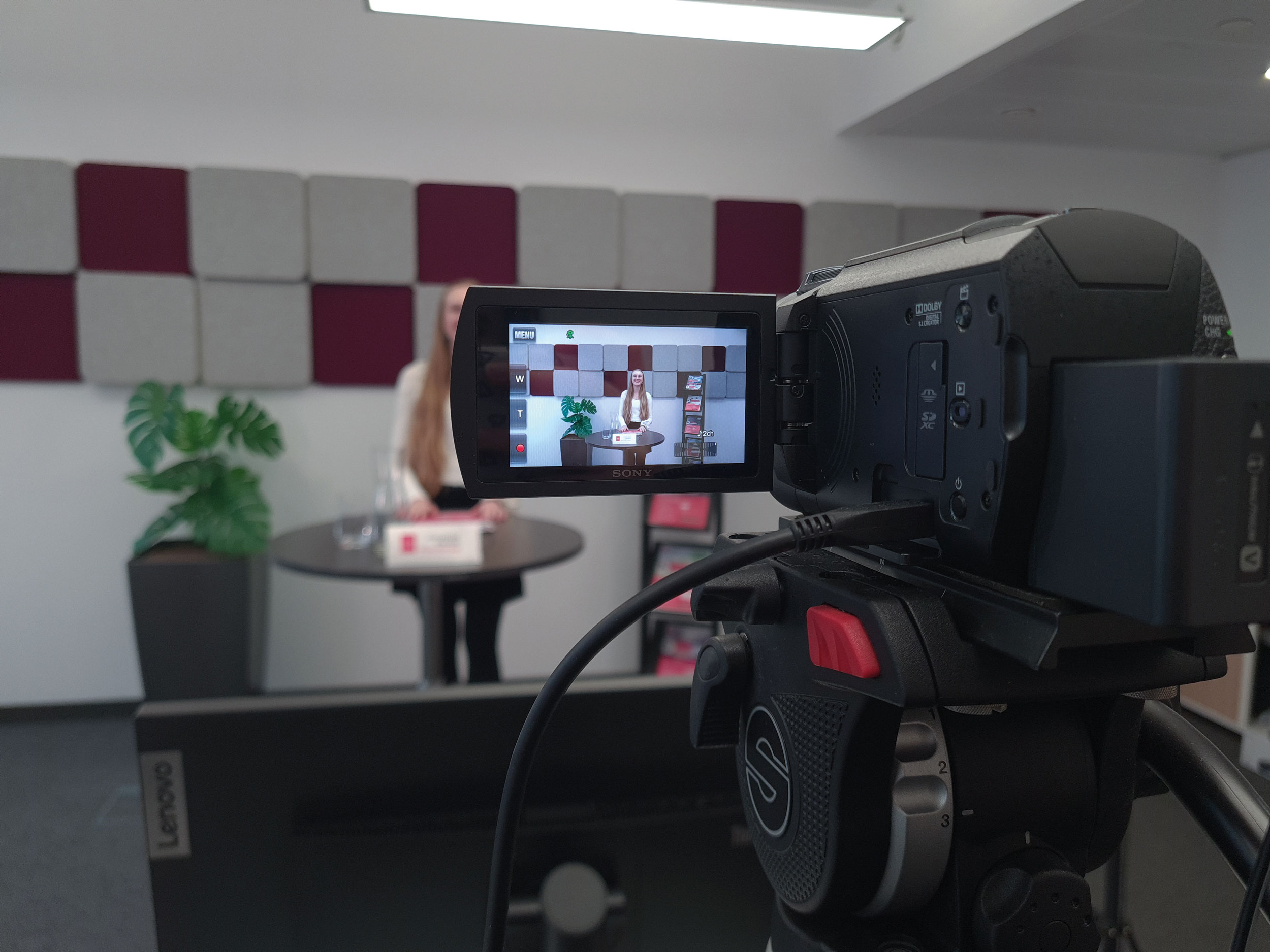 Im Vordergrund ist eine Filmkamera, durch den Bildschirm sieht man eine Frau, die bei einem Stehtisch steht und vorträgt.
