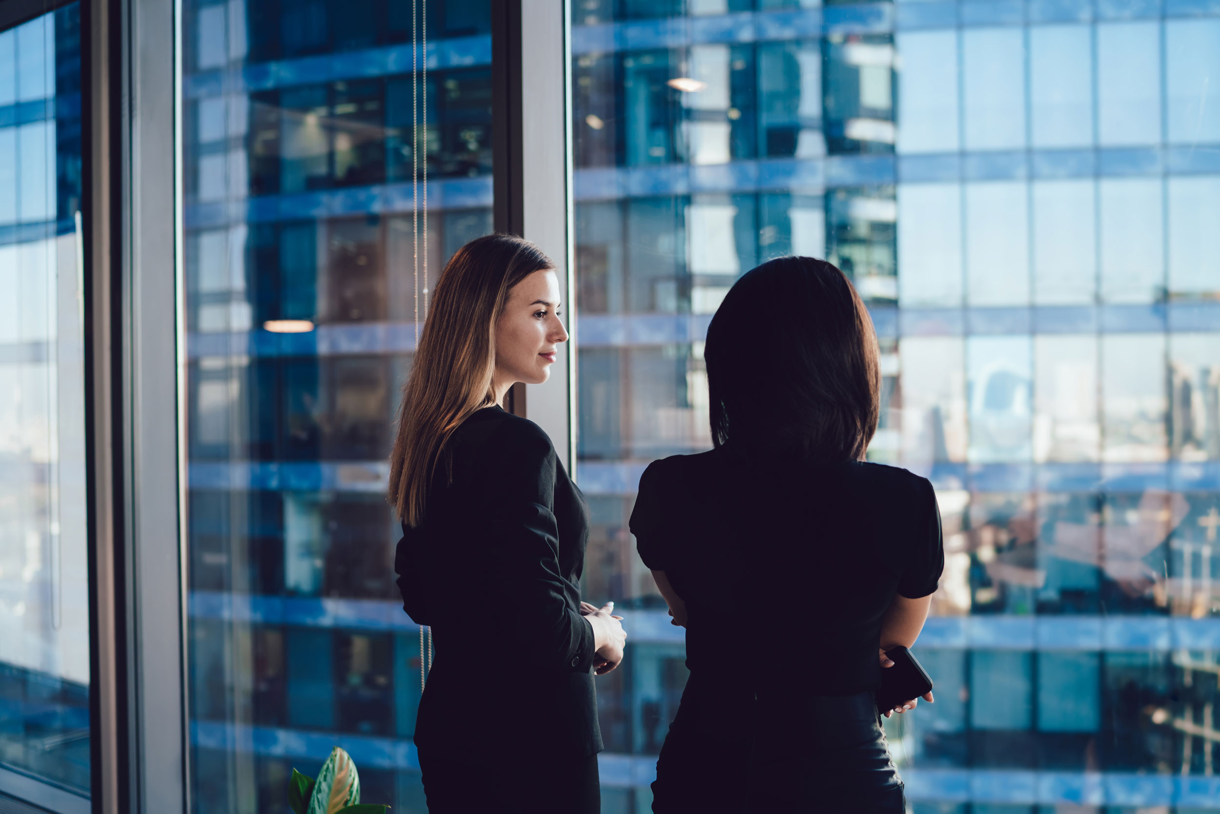 2 Frauen vor einer Fensterfront. Durchs Fenster sieht man ein Bürogebäude.