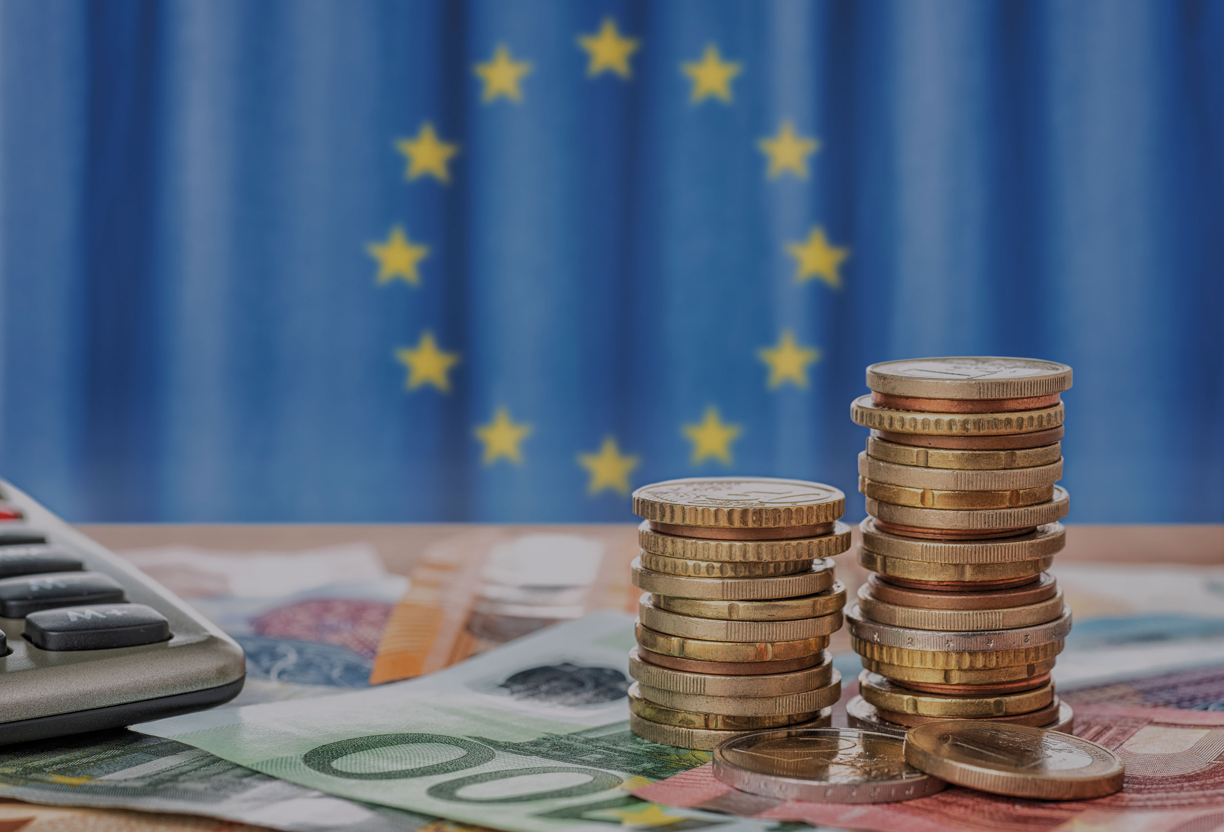 Euromünzstapel und -Banknoten, sowie ein Tacshenrechner liegen auf einem Tisch. Im hintergrund ist eine EU Flagge.