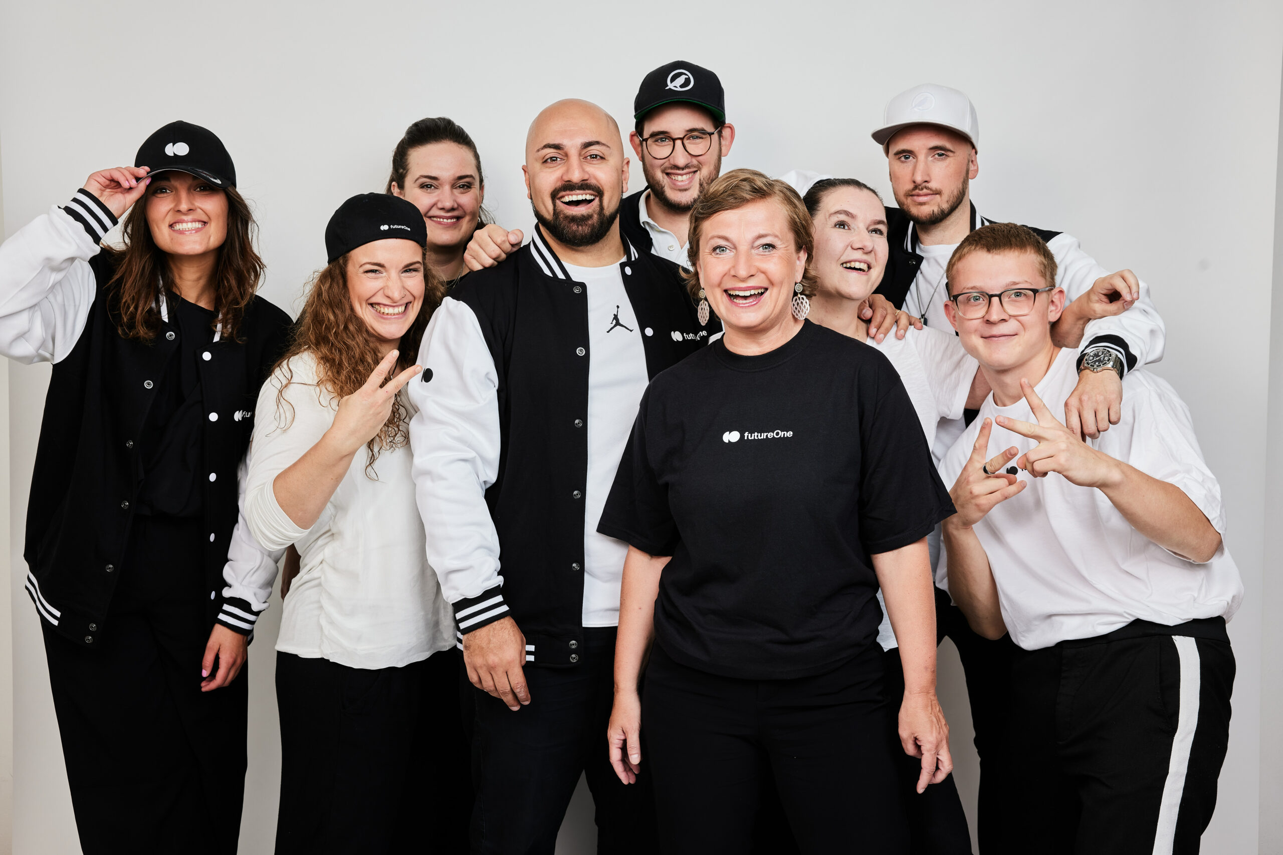 Das Team des österreichisch-persischen Keynote Speakers Ali Mahlodji steht vor einem grauen Hintergrund. Ali Mahlodji ist in der Mitte zu sehen, um ihn herum mehrere Personen in schwarz-weißer Kleidung, die in die Kamera lächeln.